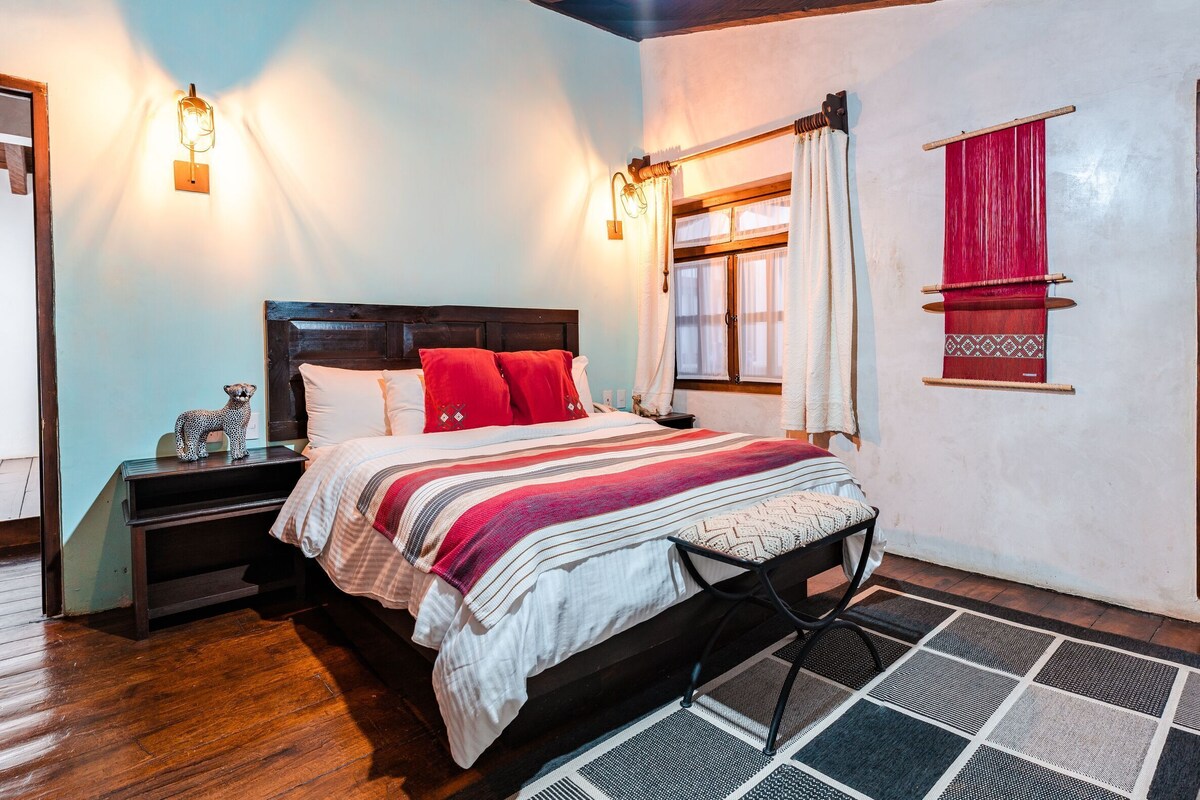 A bedroom with a wooden bed, bedside tables, a nightstand, a window with curtains, and a view of the outside.