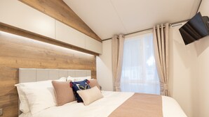 A hotel room with a wooden headboard, a large bed with multiple pillows, a TV mounted on the wall, and a window with curtains. - The Harris Hut - a fantastic new property on Sandhills Holiday Park, Mudeford (Christchurch)