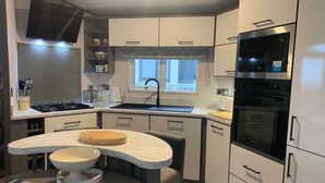 A modern kitchen with a built-in stove, oven, and microwave. There is a window above the sink and a small island with a cutting board and bowl. - The Harris Hut - a fantastic new property on Sandhills Holiday Park, Mudeford (Christchurch)