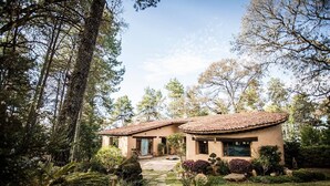 A two-story house with a tiled roof, surrounded by a lush garden and tall trees.