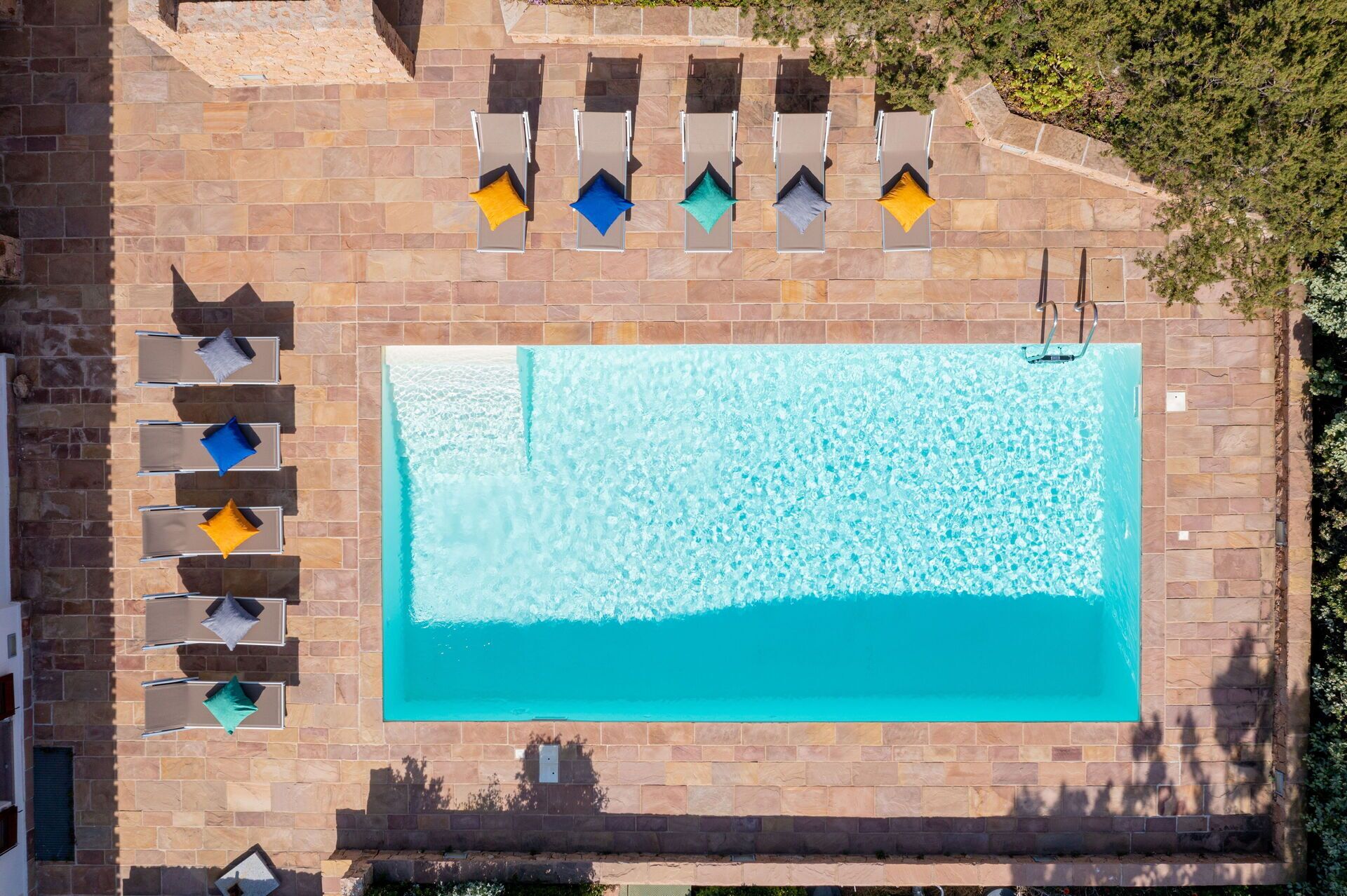 Vista aérea de una piscina rectangular con una terraza de baldosas a su alrededor, que incluye varias sillas de descanso con cojines.