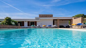 A swimming pool with lounge chairs and umbrellas, a stone wall, and a building with a tiled roof. - Villa Belle: private pool near the beach (Aglientu)