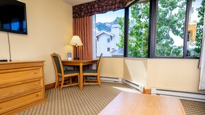 Een hotelkamer met een houten dressoir, een tafel, twee stoelen, een aan de muur bevestigde televisie en een raam met uitzicht op gebouwen en bomen.