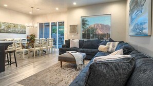 A modern living room with a dark gray sofa, a dining area with white chairs, and a large window with blinds.