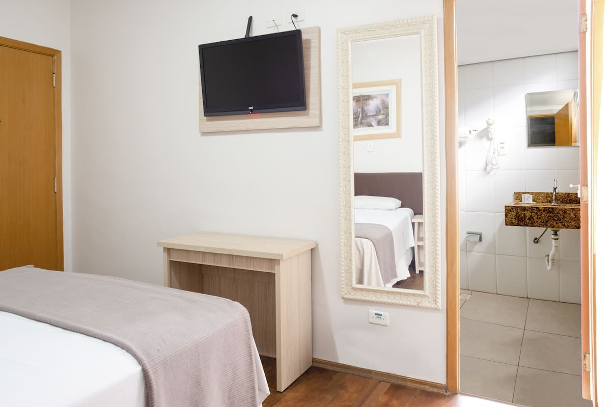 A bedroom with a bed, a TV mounted on the wall, a wooden desk, and a bathroom visible through an open door.