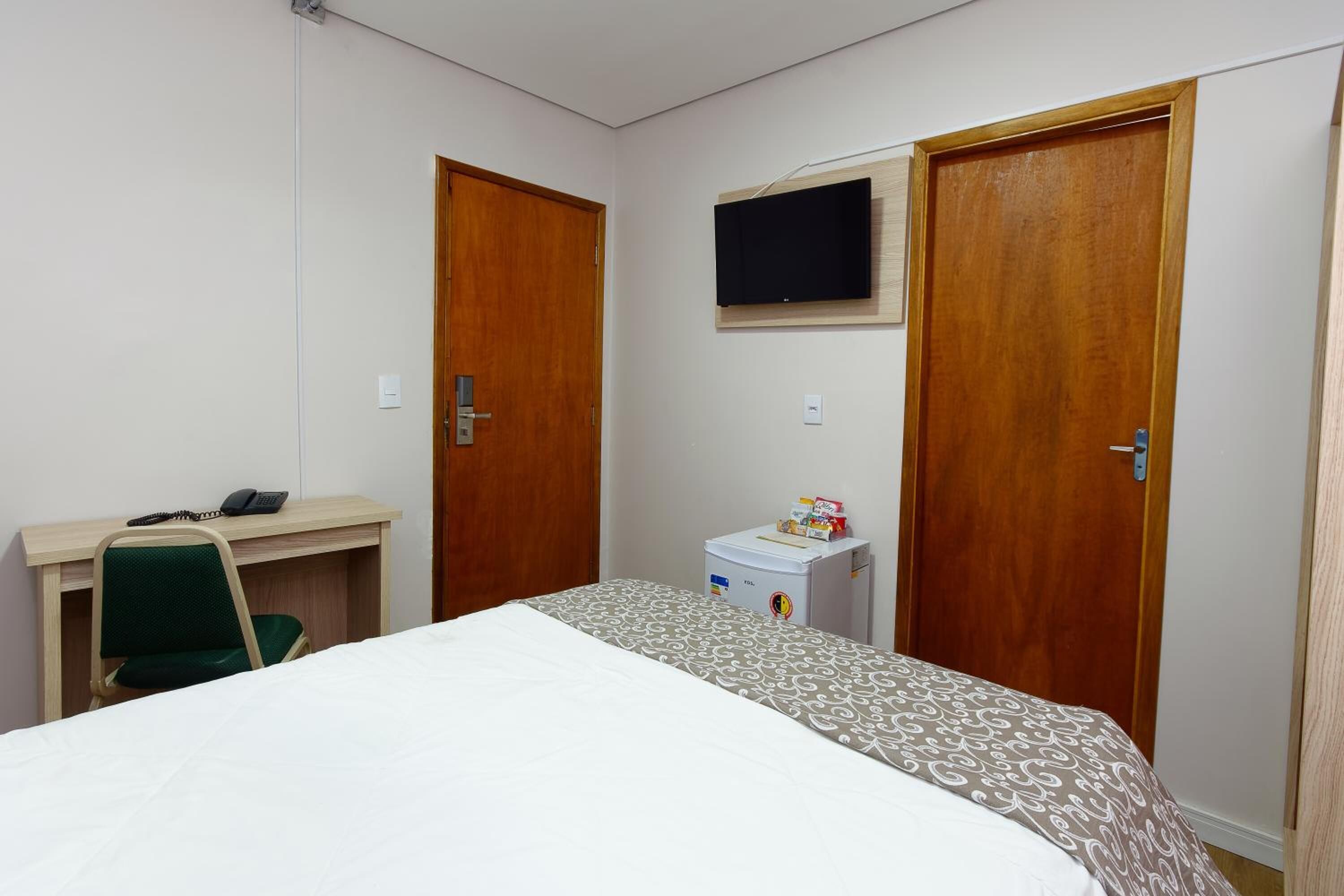 Una habitación de hotel con cama, escritorio con teléfono, televisor montado en la pared y un refrigerador pequeño.