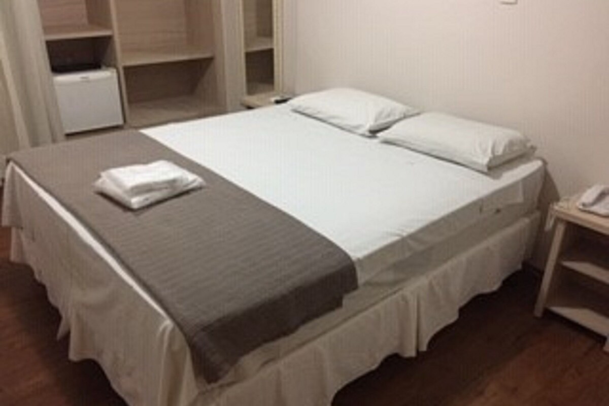 A neatly made bed with a white comforter and pillows, a gray throw blanket, and a white towel on the side.