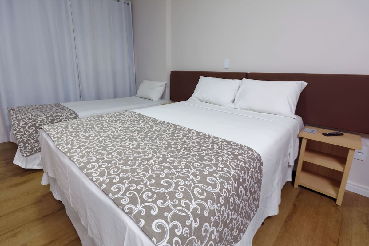 A hotel room with two beds, each with a patterned bedspread, a wooden nightstand with a remote control, and a white curtain.