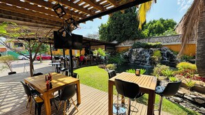 An outdoor patio with wooden tables and chairs, a TV screen, and a garden with a pond and waterfall. - Hotel Villa Pralaya (Jiutepec)
