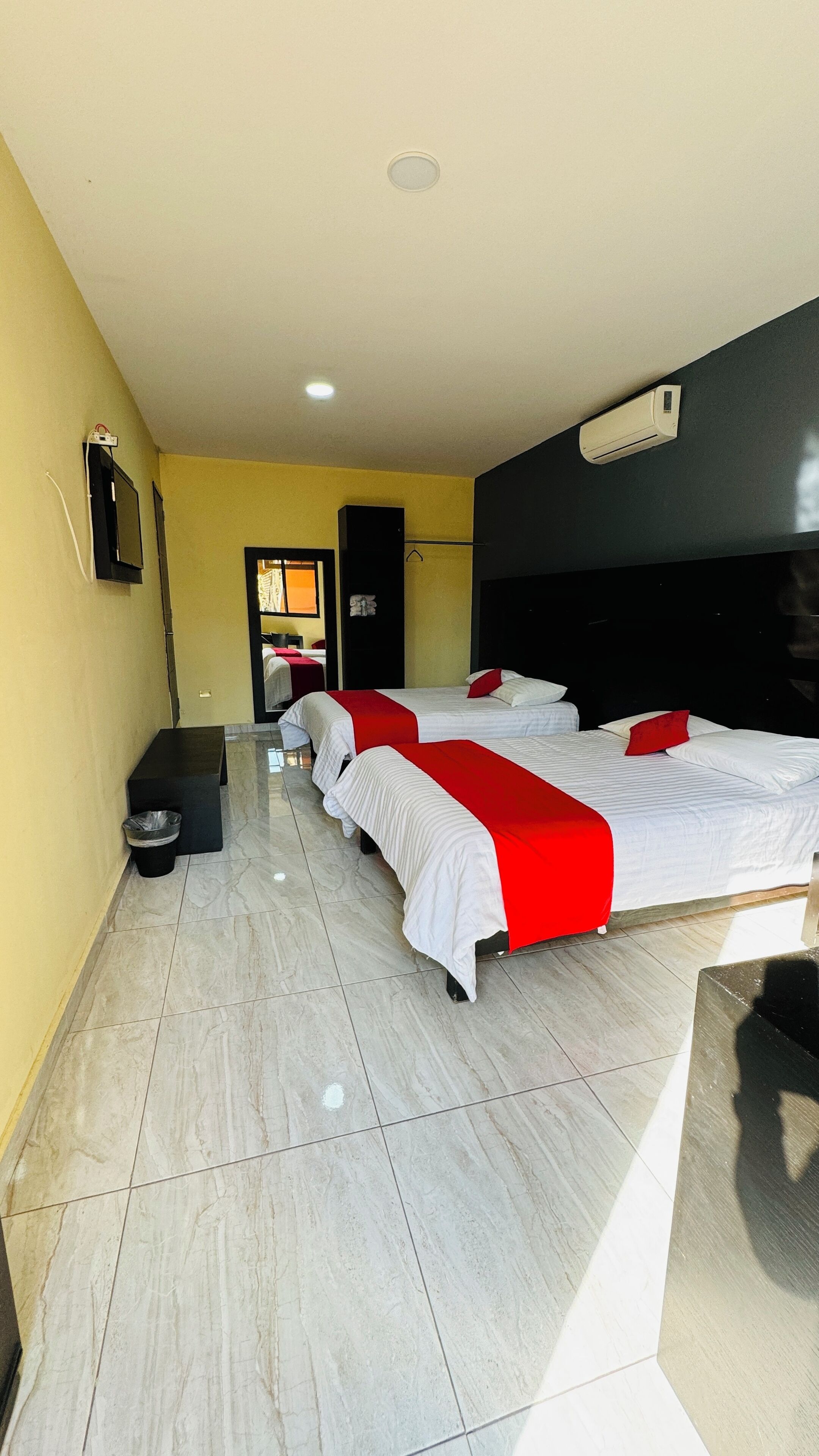 A hotel room with two beds, a black headboard, a ceiling light, and a wall-mounted air conditioner.