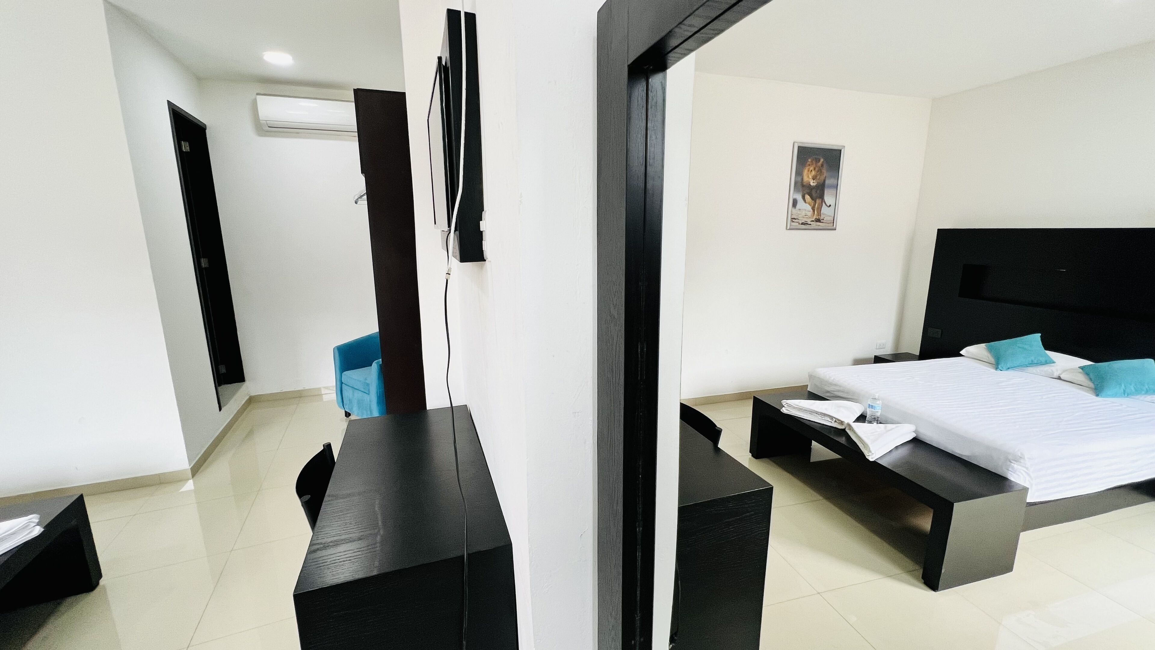 Une chambre d’hôtel moderne avec un grand lit, un bureau et une chaise. Un tableau est accroché au mur et un appareil de climatisation est installé au-dessus de la porte.