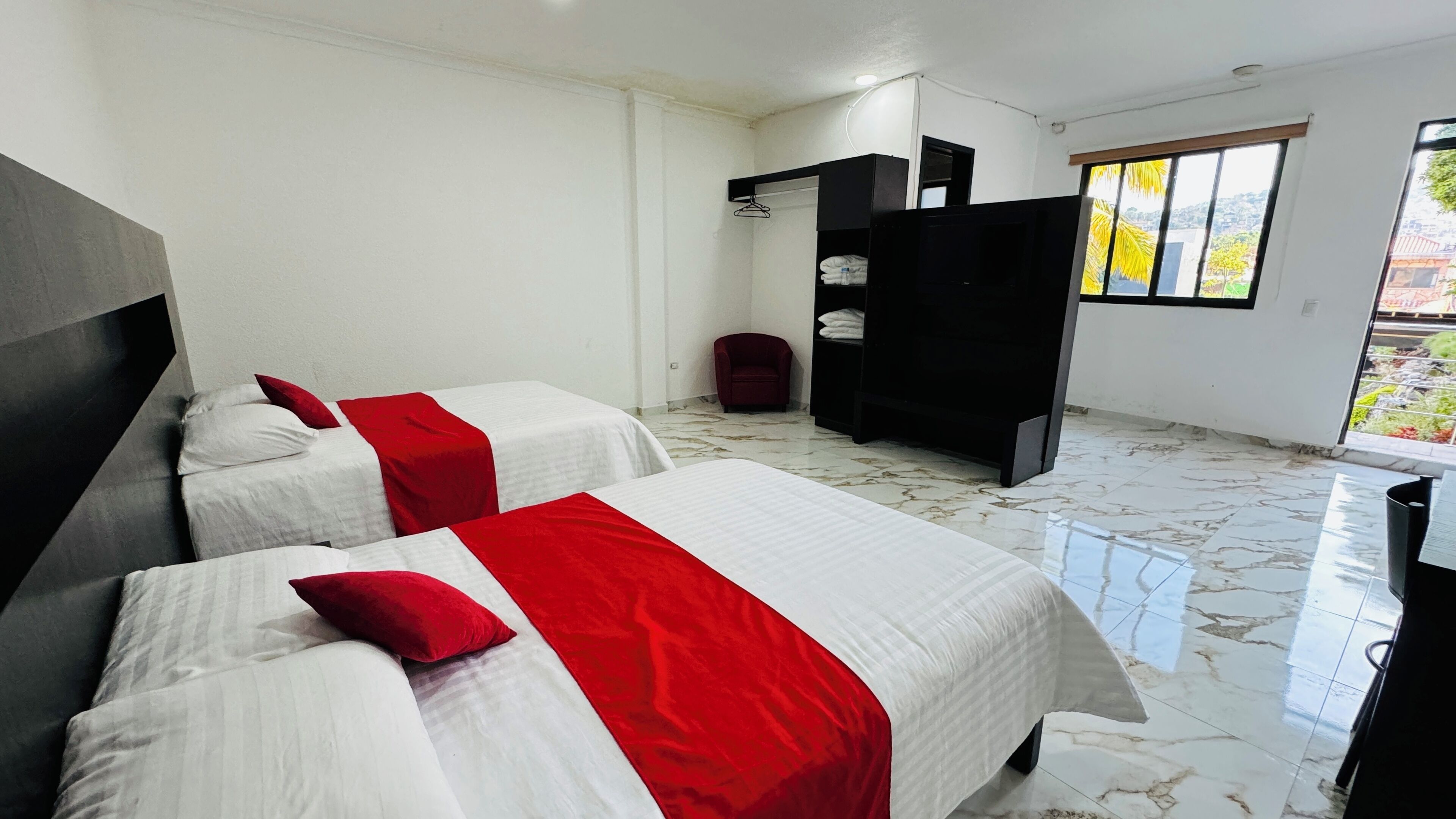 A hotel room with two beds, a wardrobe, a chair, and a view of greenery outside.