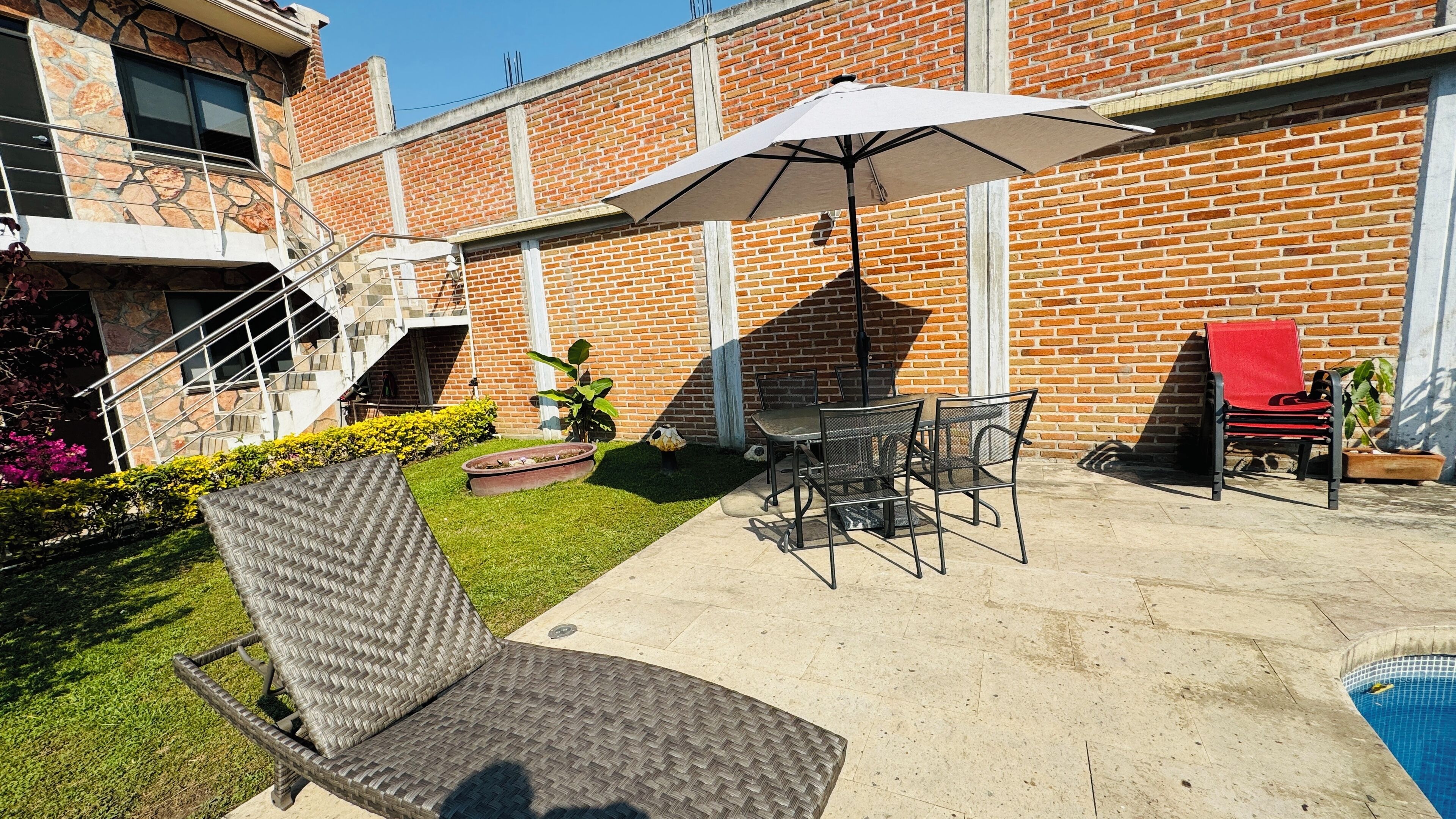 A patio area with a dining table, chairs, a wicker lounge chair, a white umbrella, and a swimming pool.