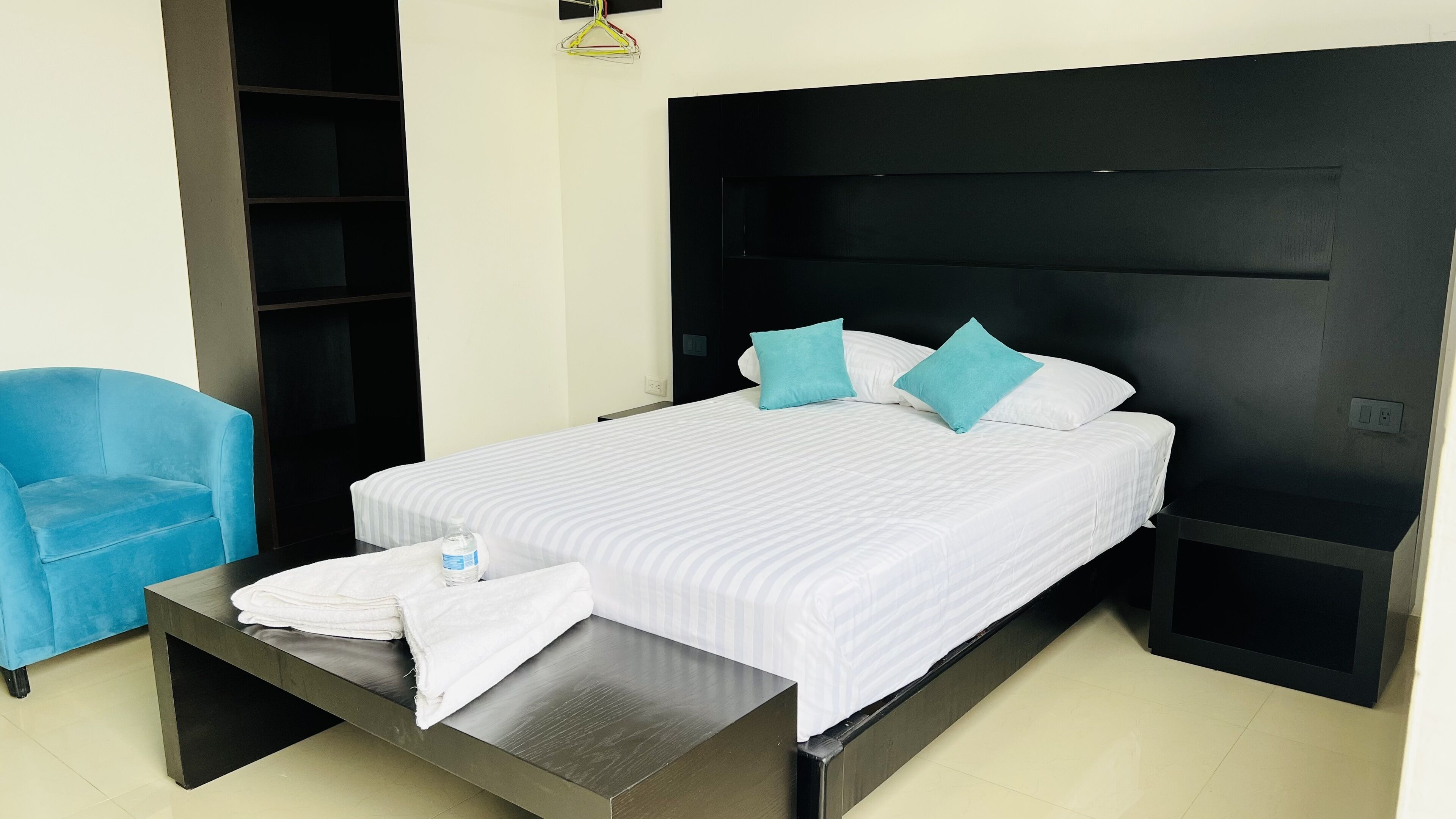 A hotel room with a bed, a blue chair, a bedside table with a bottle and towels, and a black headboard.