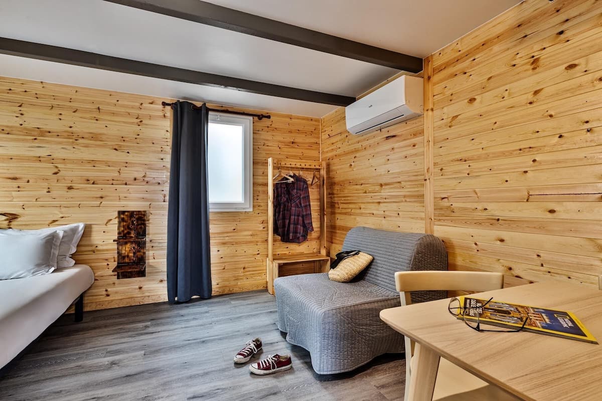 Ein gemütliches Zimmer mit Holzwänden, einem Bett, einem grauen Sofa, einem Tisch mit einer Zeitschrift und einem an der Wand befestigten Kleiderhaken.