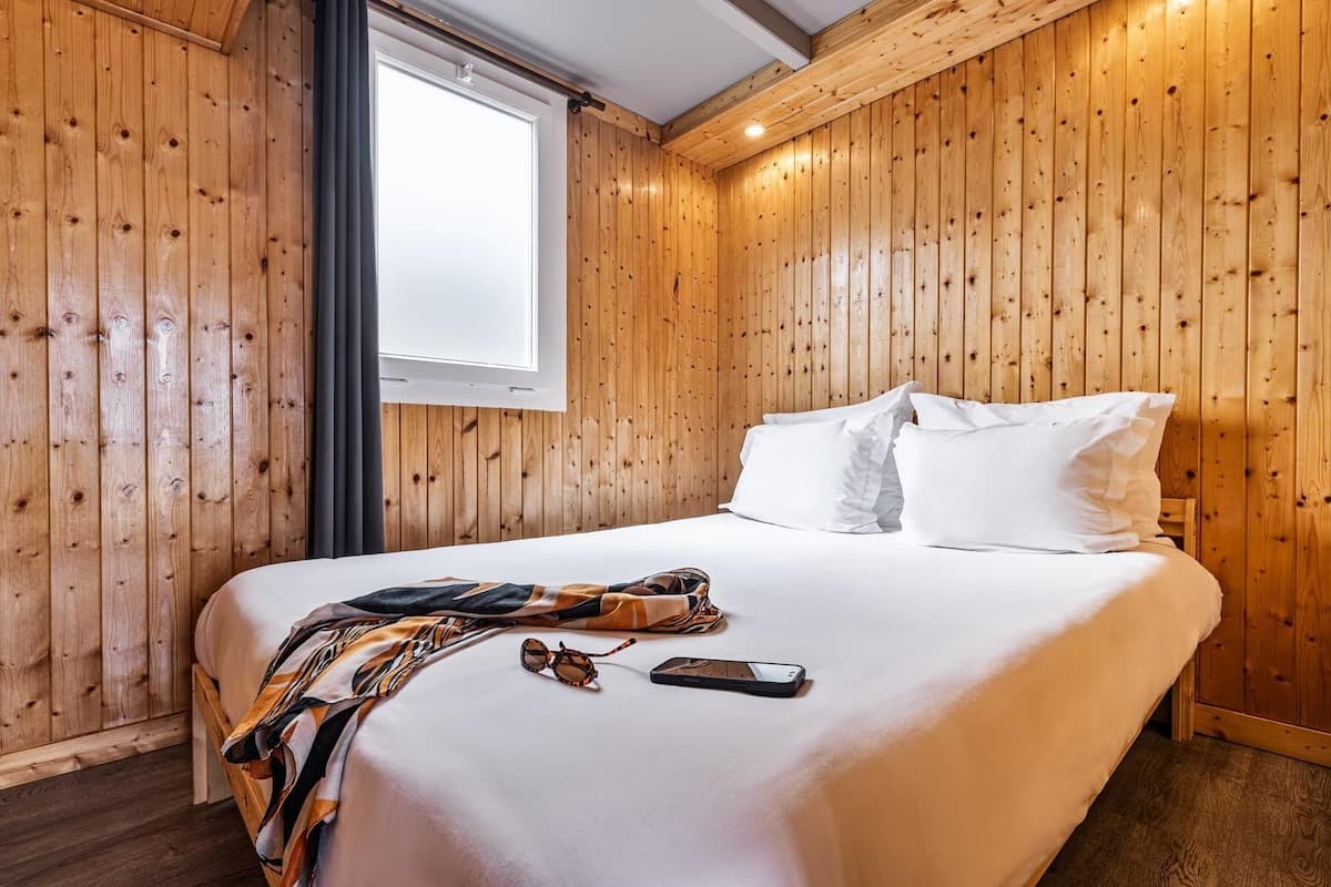 Ein Zimmer mit Holzwänden, einem Bett, einem Fenster und Blick ins Freie.