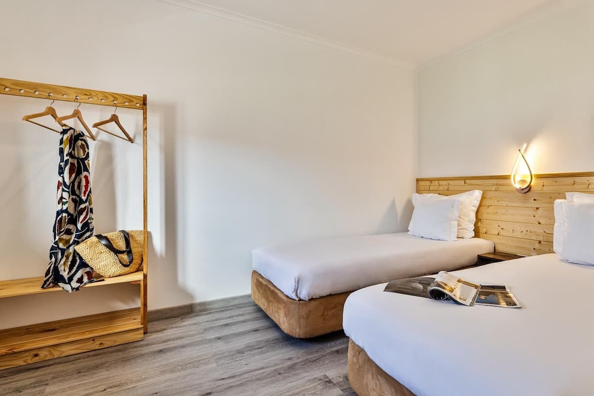 Ein Hotelzimmer mit zwei Betten, einem Holzkopfende, einem Holz-Kleiderständer mit einem gemusterten Kleidungsstück und einer Zeitschrift auf dem Bett.