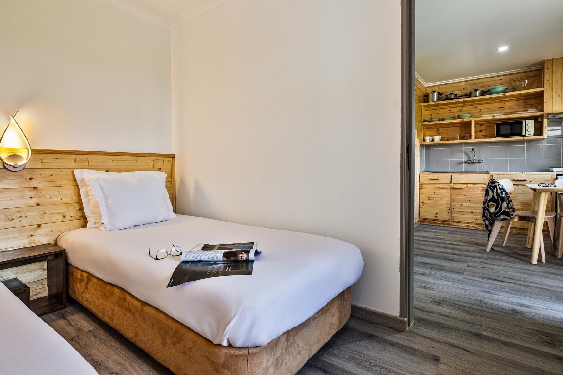 Ein Hotelzimmer mit einem Holzkopfteil, einem Bett mit weißer Bettwäsche, einem Nachttisch mit Gläsern und einer Zeitschrift sowie Blick in den angrenzenden Küchenbereich mit Holzschränken und einem Esstisch.