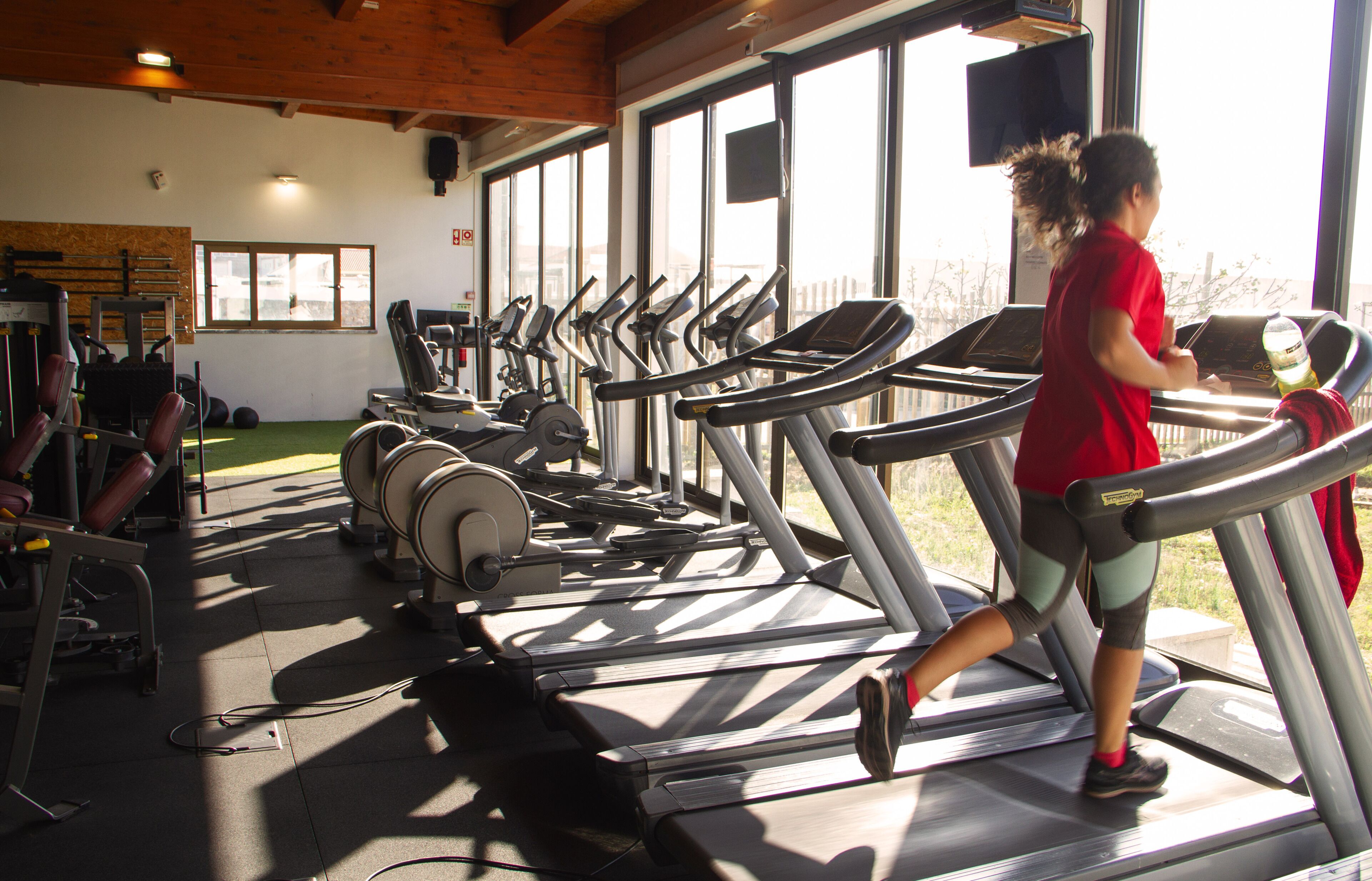 Un gimnasio con numerosas cintas de correr, máquinas de pesas y un amplio ventanal que proporciona luz natural.