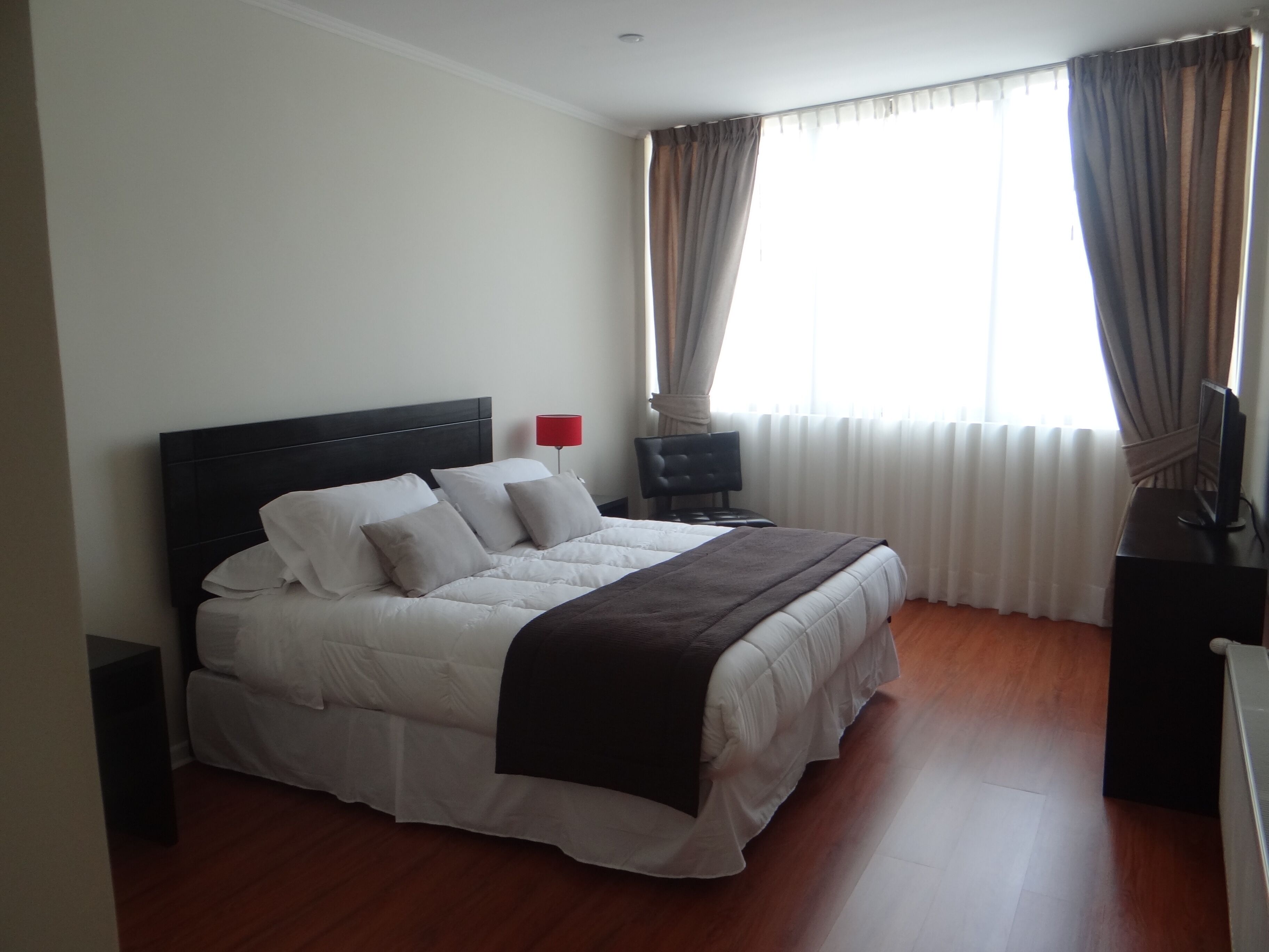A neatly made bed with white bedding and a dark headboard in a room with wooden flooring.