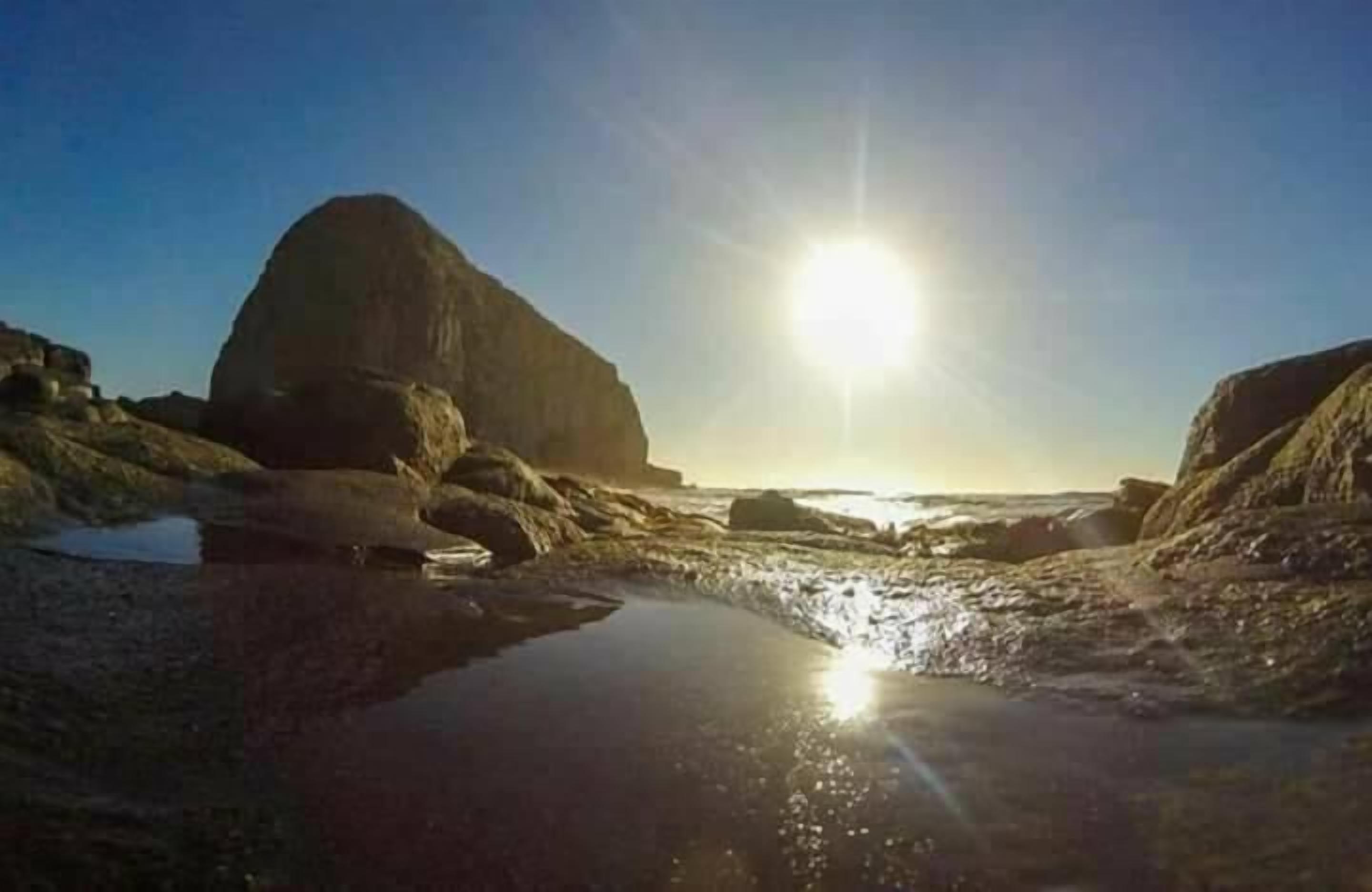 A rocky beach with a large boulder and a small pool of water reflecting the sun.