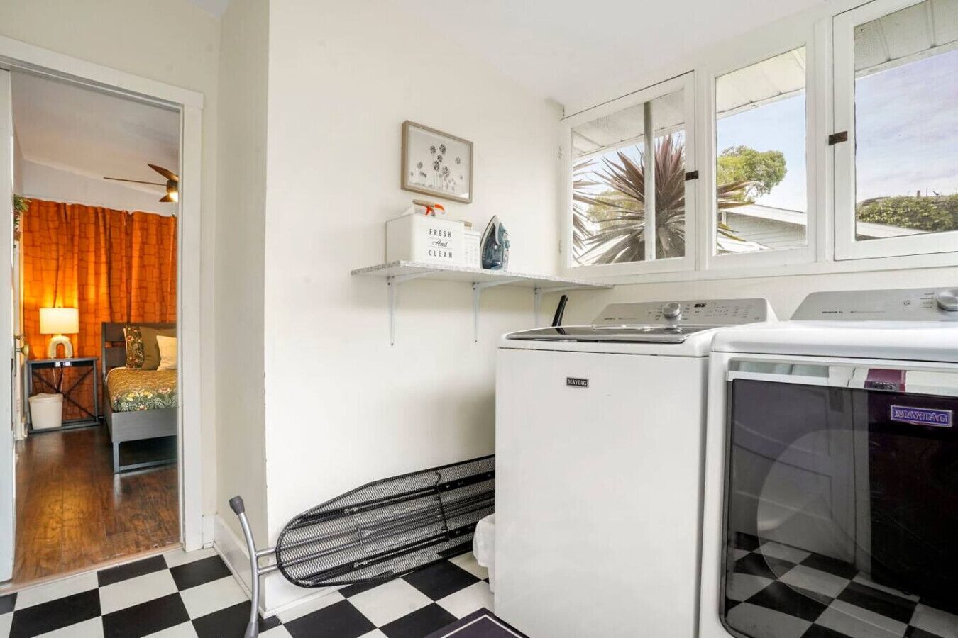 A compact hotel room with a checkered floor, a bed, a washing machine, and a window with a view of palm trees.