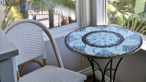 Een kamer met een rieten stoel, een klein rond tafeltje met een mozaïek blad, en uitzicht op palmbomen door een raam.