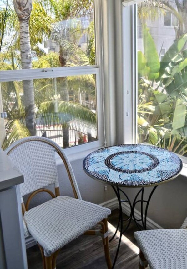 A room with a wicker chair, a small round table with a mosaic top, and a view of palm trees through a window.