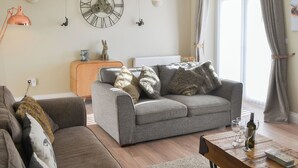 Sala de estar com sofá cinzento, mesa de centro de madeira com copos de vinho, relógio na parede e janela com cortinas.