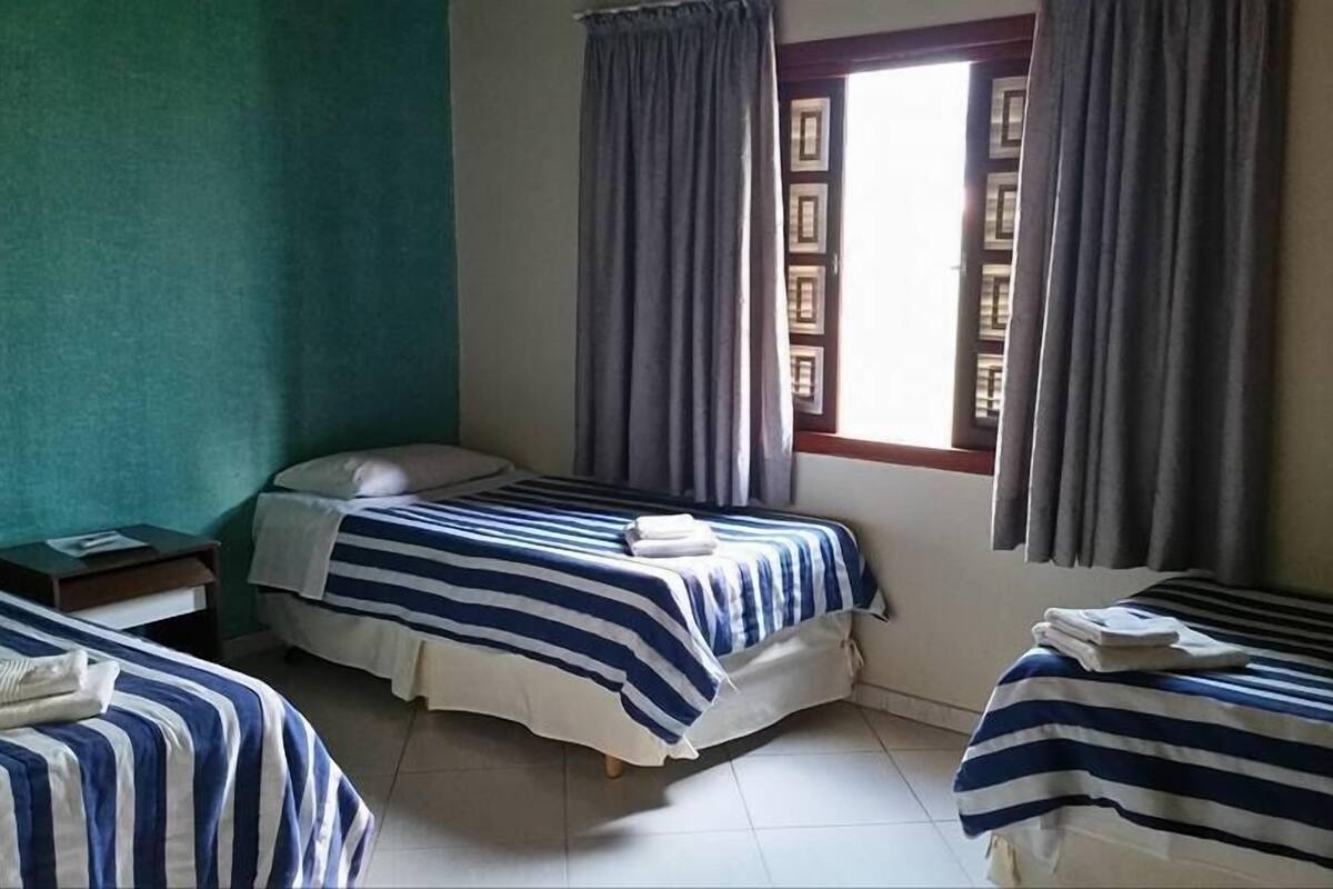 A room with two beds, each with striped bedding, a small bedside table, and a window with curtains.