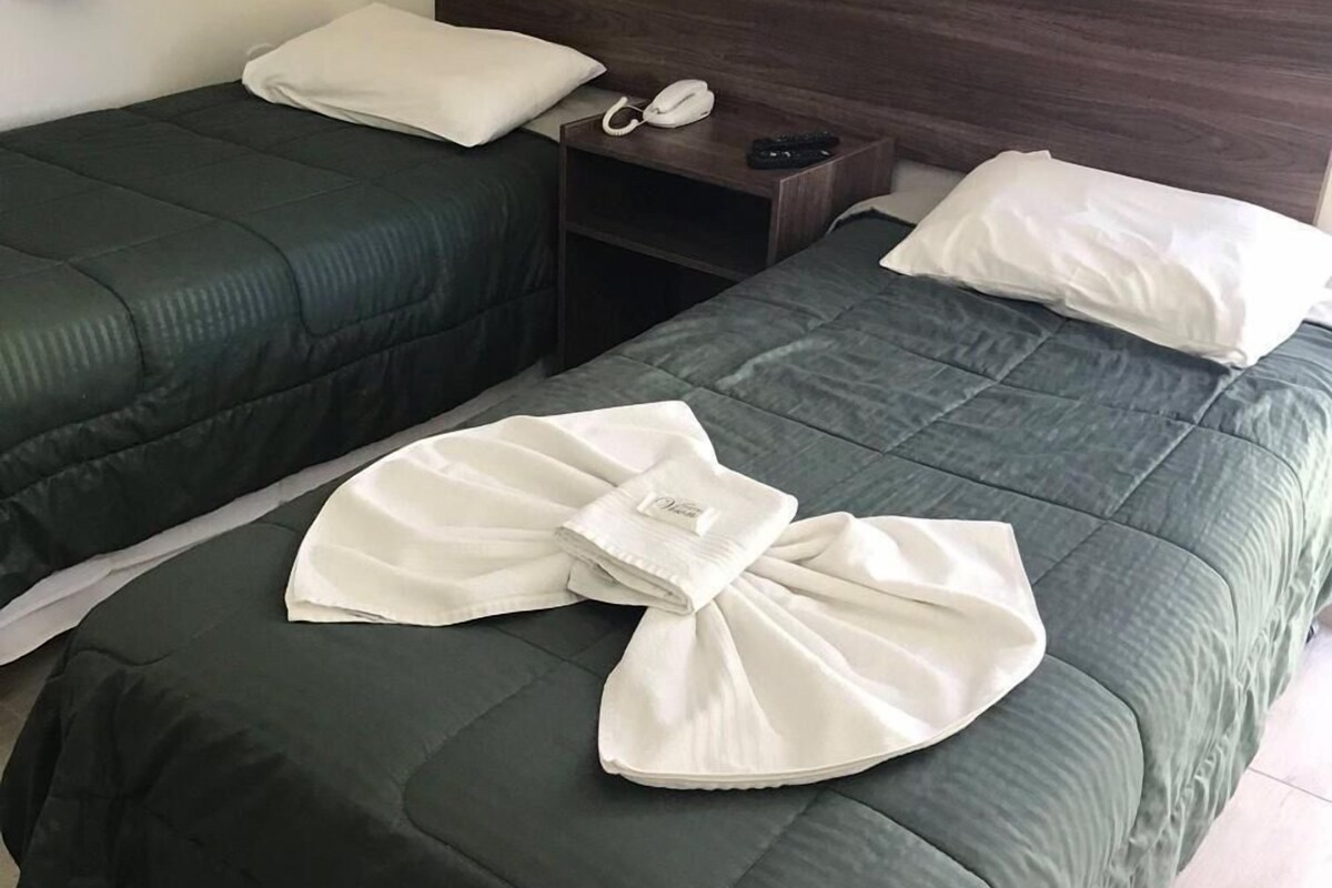A hotel room with two single beds, each with a green bedspread and white pillows. There is a wooden headboard, a bedside table with a phone and a remote control, and a folded white towel on the bed.