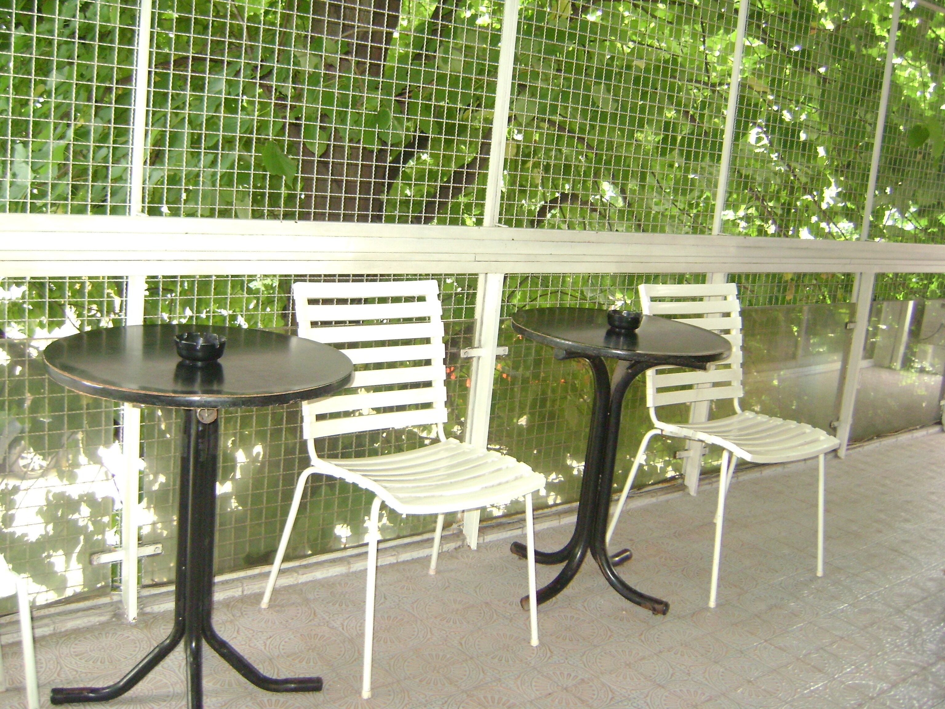 Two white metal chairs and tables with black metal legs, positioned in front of a glass window with a green leafy background.