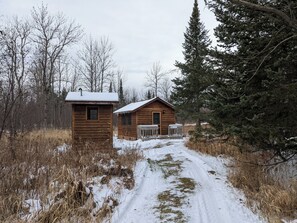 Due cabine di legno con un sentiero innevato che le conduce, circondate da alberi e erba secca.