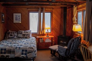 A wooden cabin room with a bed, a chair, a table with a lamp, and a wood-burning stove.