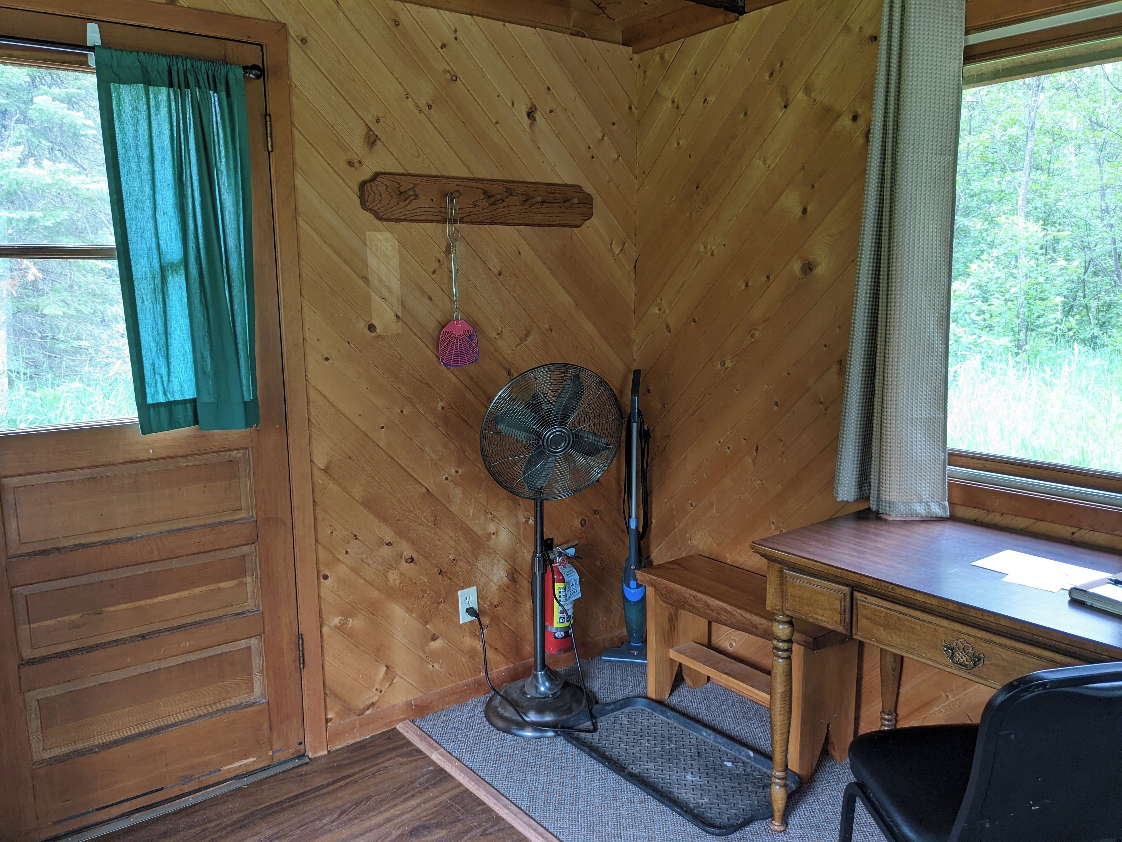 A wooden cabin interior with a desk, chair, fan, and a window with curtains.