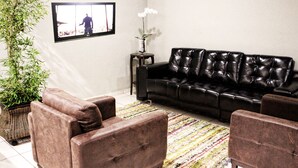 Una sala de estar con un sofá de cuero negro, dos sillones marrones, un tapete con rayas de colores, una planta en maceta y una mesita con un jarrón.