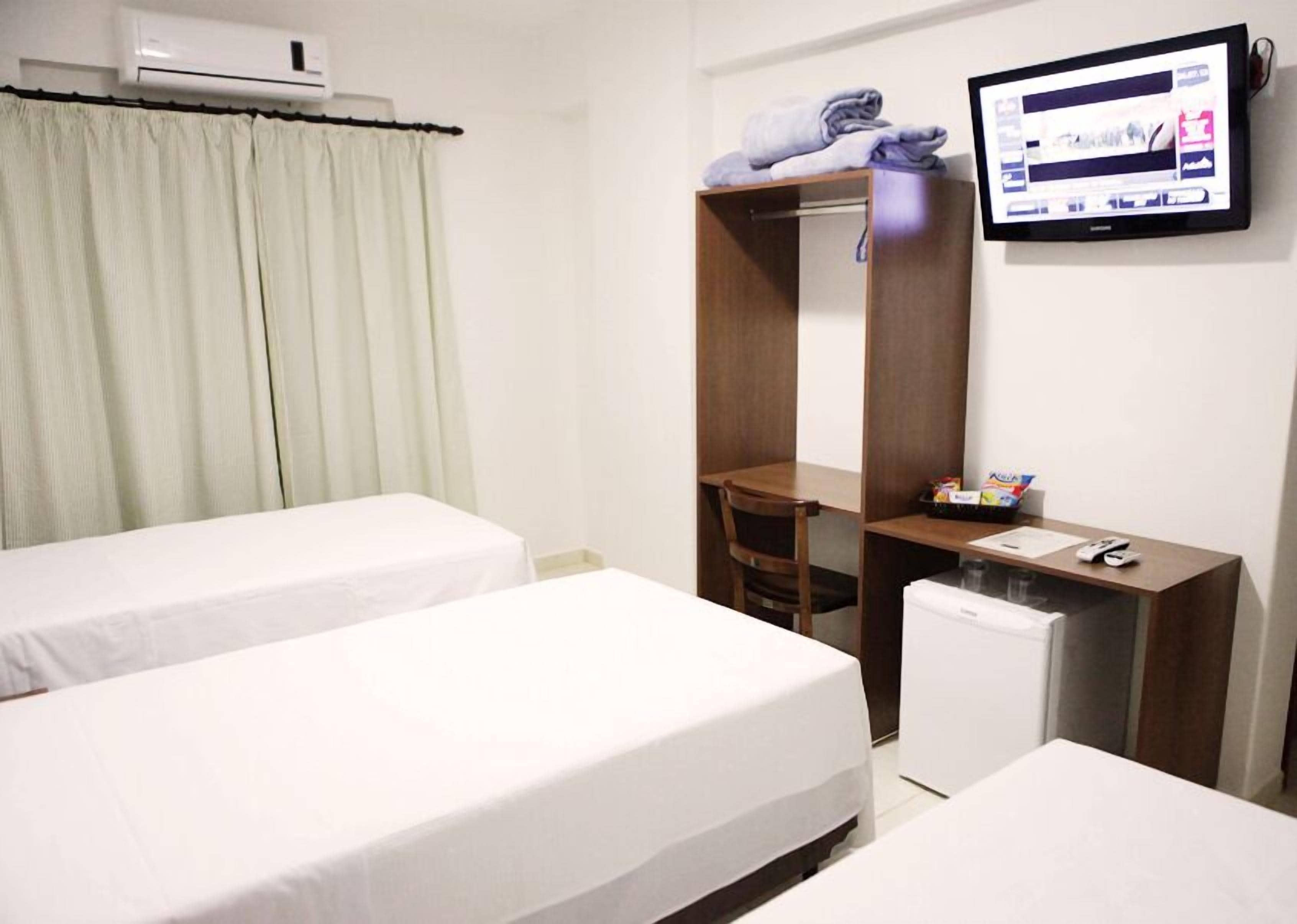 Habitación de hotel con dos camas individuales, un escritorio con silla, un refrigerador pequeño y un televisor montado en la pared.
