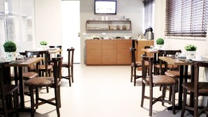 Un comedor moderno con mesas y sillas de madera, una zona de cocina con un armario de madera, y un televisor montado en la pared.