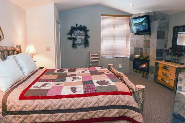 A bedroom with a large bed, a fireplace, a television, and a window with blinds. - Fawn Valley Inn: 103 (Estes Park)