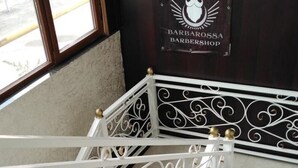 Un escalier en métal blanc, orné de motifs complexes, mène à une pièce dont un mur est recouvert de bois sombre et sur laquelle on peut lire l’inscription « Barbossa, salon de coiffure ».