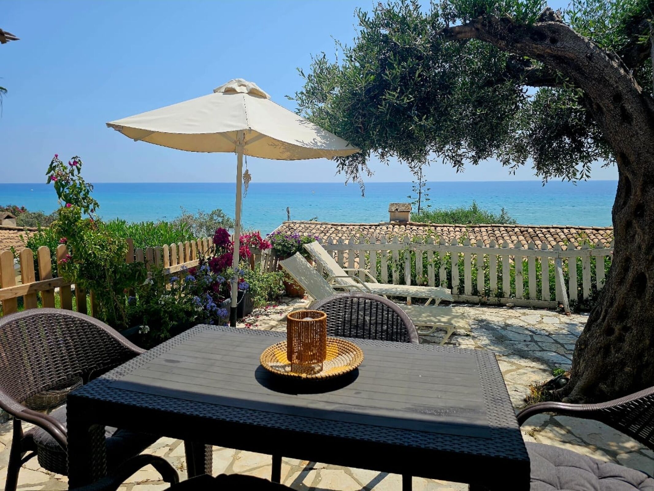 A patio with a table and chairs, a white umbrella, and a view of the sea.