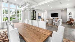 Een ruime woonkamer met een grote houten eettafel, omringd door witte fauteuils, een open haard en een aan de muur bevestigde televisie.