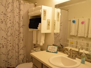 A bathroom with a floral shower curtain, a towel rack with folded towels, a sink with a mirror, and a motivational sign on the wall.