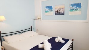 Cama bem arrumada com lençol branco e manta azul, dois rolos de toalha sobre a cama, abajur e três quadros de parede com frases motivacionais.