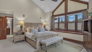 A bedroom with a large bed, two bedside tables with lamps, a bench, a window with a view, and a ceiling fan.