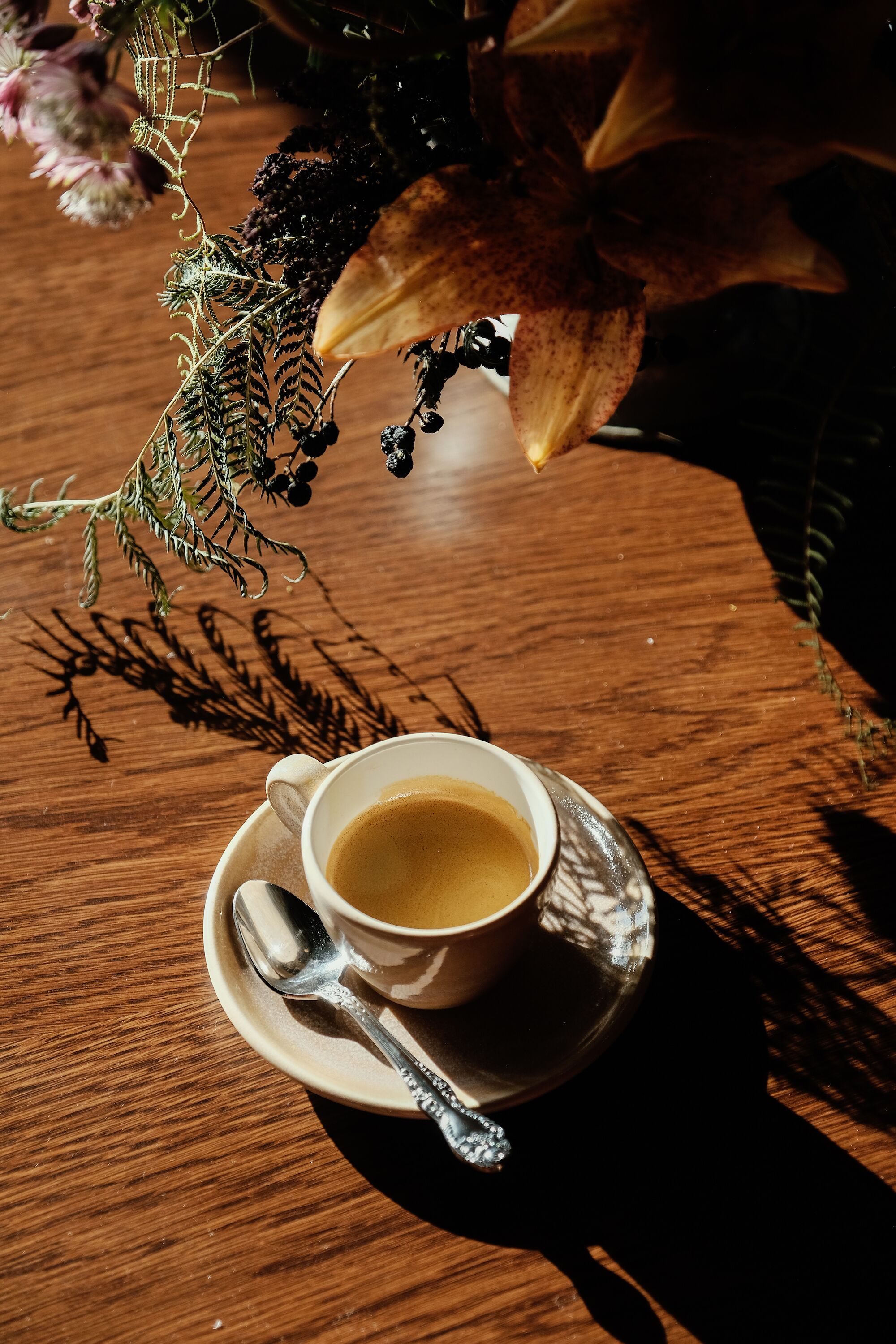 A cup of coffee on a saucer with a spoon, placed on a wooden table with shadows of leaves and branches.