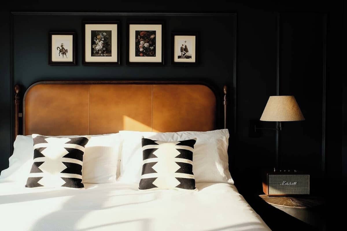 A bed with white linens and patterned pillows, a wooden headboard, and a lamp on a bedside table.