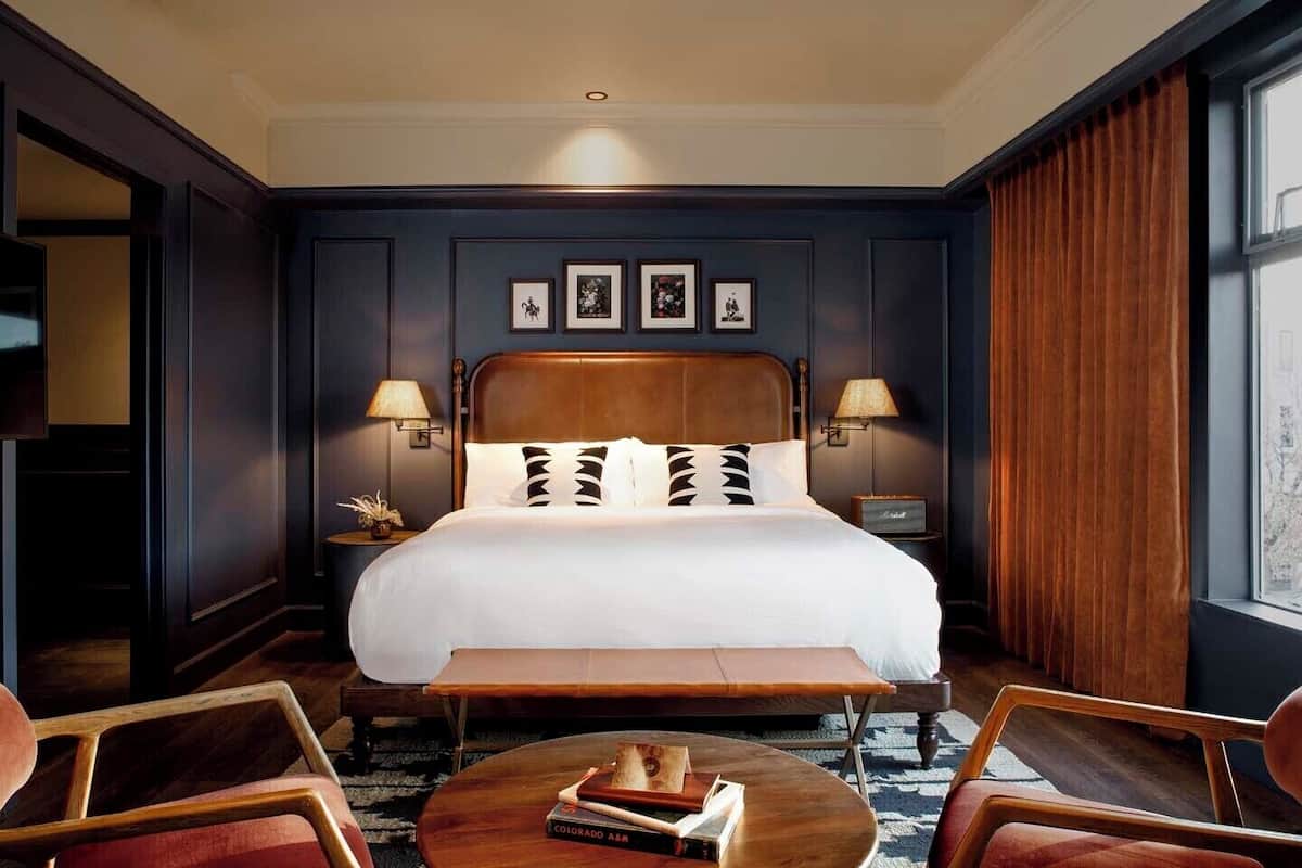A hotel room with a large bed, two bedside lamps, a wooden headboard, a round coffee table with books, and a view through a window.