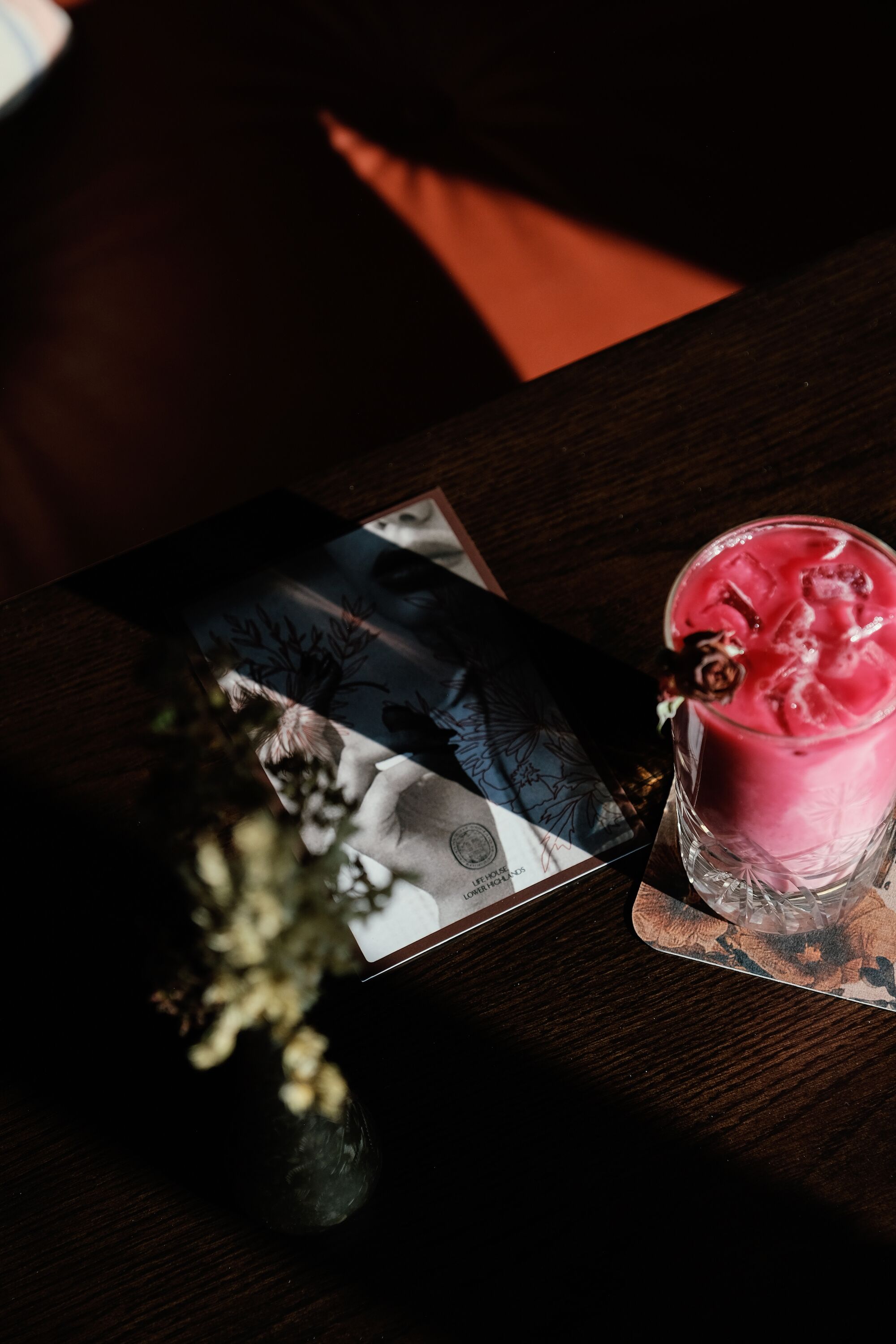 A dark wooden table with a glass of pink drink, a card, and a small vase with dried flowers.