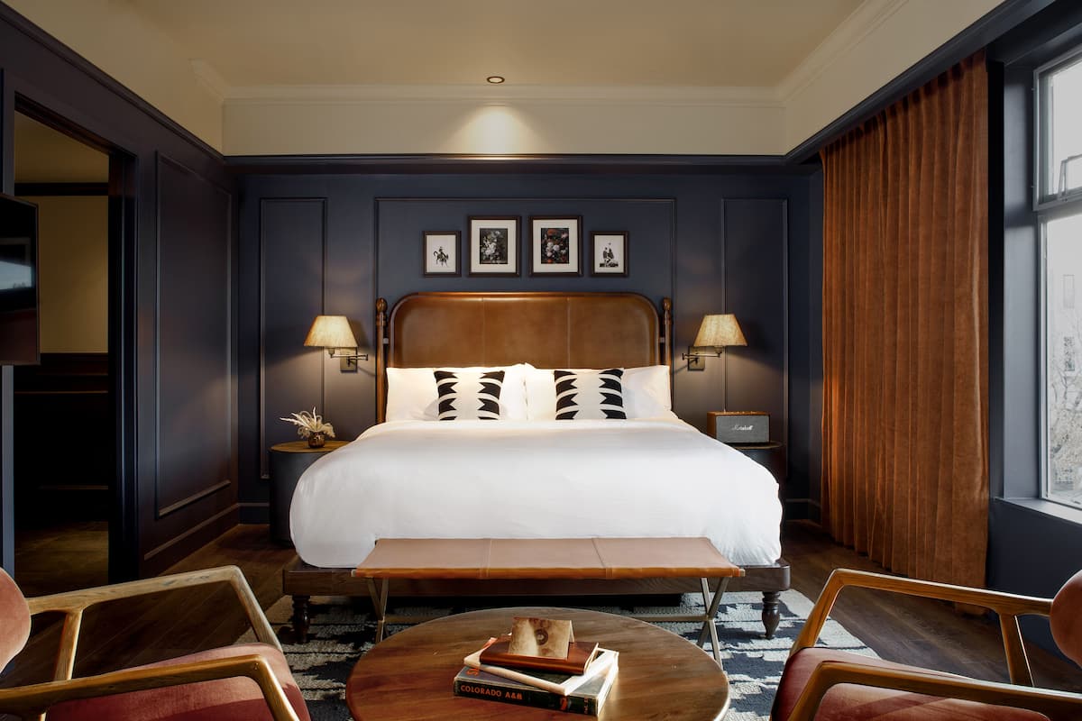 A hotel room with a large bed, two bedside lamps, a wooden bench, a round coffee table with books, and a view of the city through a window.
